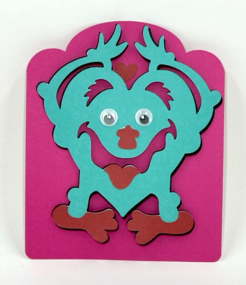 A colorful Valentine's Day card featuring a teal monster on a pink background, crafted for children.